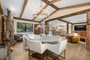 A spacious living area with a round dining table, white chairs, a chandelier, and wooden beams on the ceiling.
