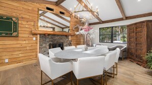 A dining area with a round table and white chairs, a chandelier, a stone fireplace, wooden walls, and a large window.