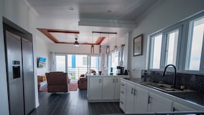 A modern kitchen with white cabinets, a black countertop, and a gas stove. There is a dining area with a brown chair and a view of the outdoors through large windows. - Majestic Dream Villas (Choiseul)