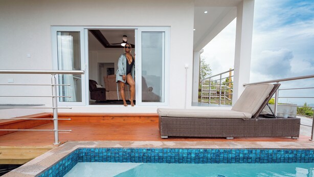 A modern outdoor pool area with a clear view of the pool, a wicker lounge chair, and a glass door leading to an interior space. - Majestic Dream Villas (Choiseul)