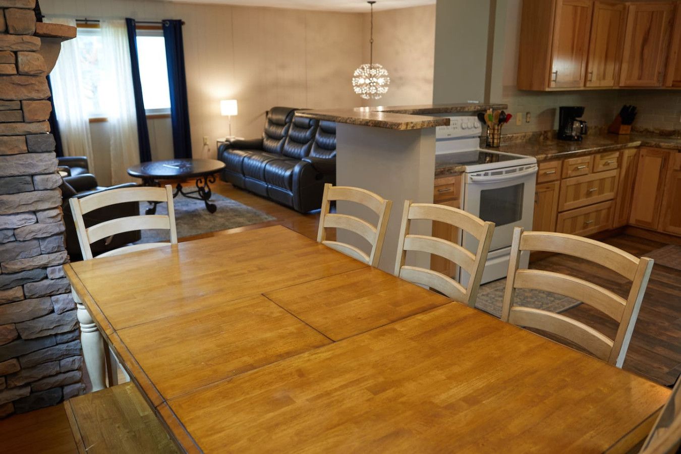 Un comedor con mesa y sillas de madera, una pared de acento de piedra, un sofá de cuero negro y una cocina con armarios de madera y una cafetera.