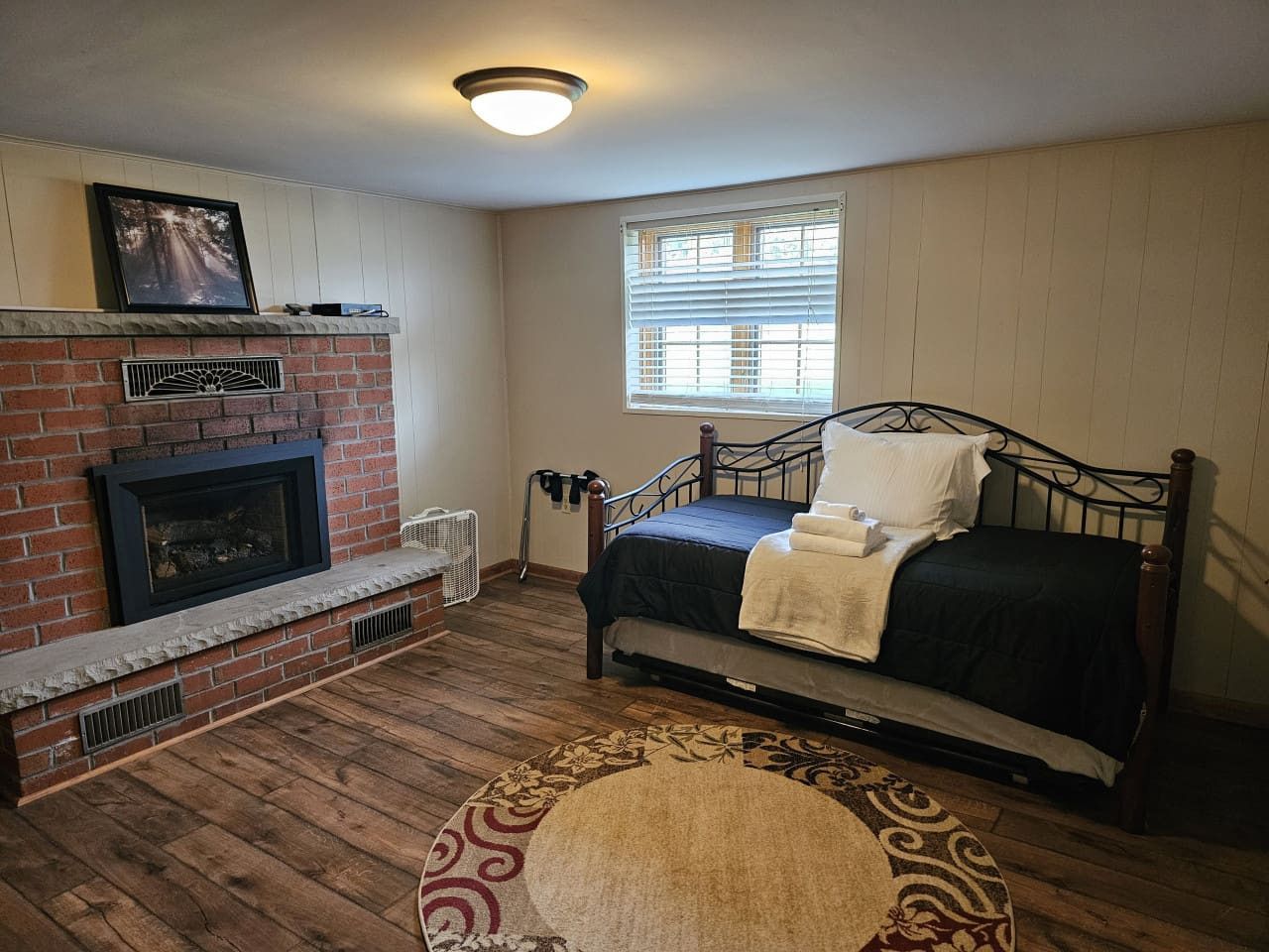 Un dormitorio con chimenea de ladrillo, una cama con colcha azul oscuro, una ventana con persianas y suelo de madera.