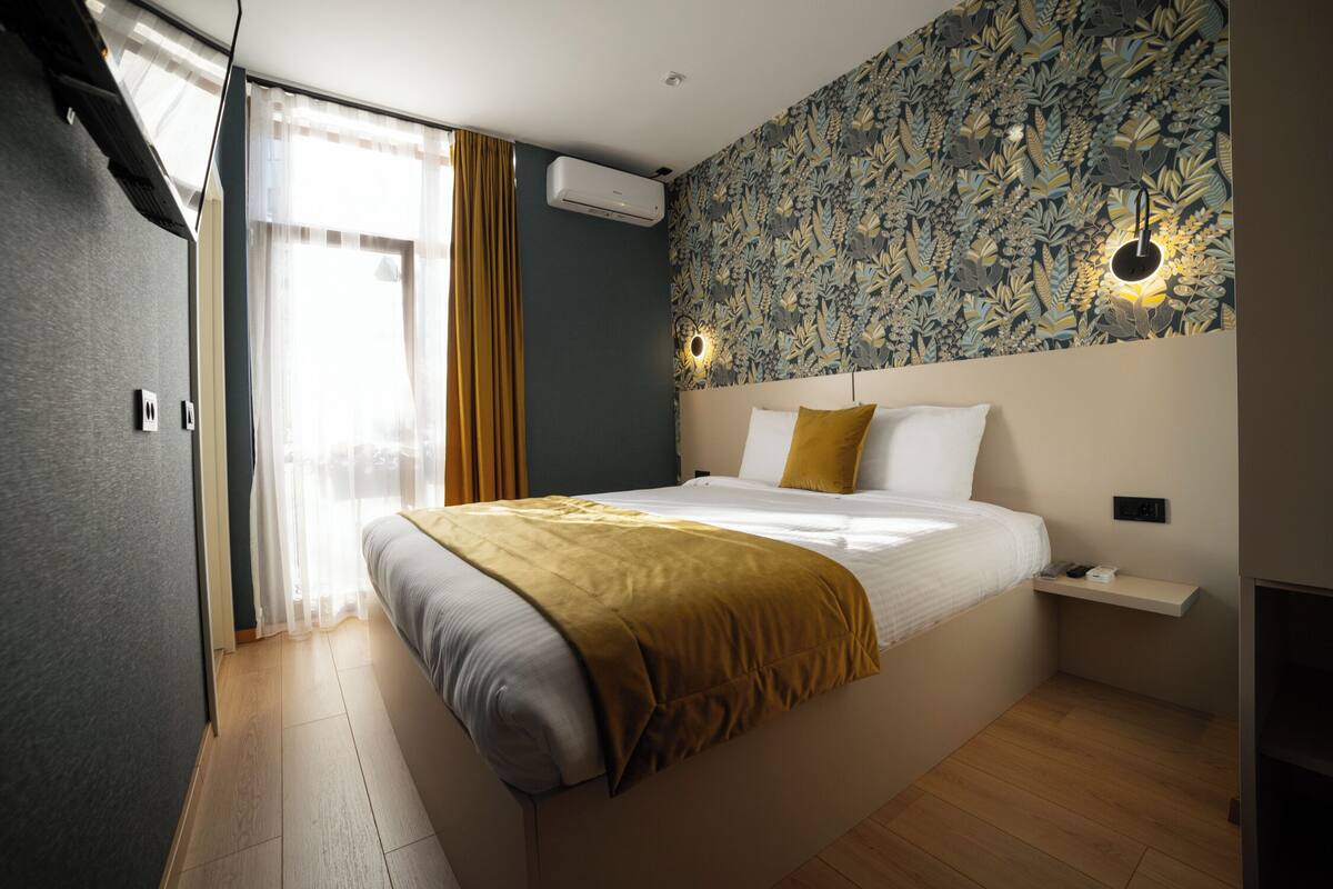 A modern bedroom with a large bed, a headboard with built-in lighting, a small bedside table, and a wall-mounted air conditioner.