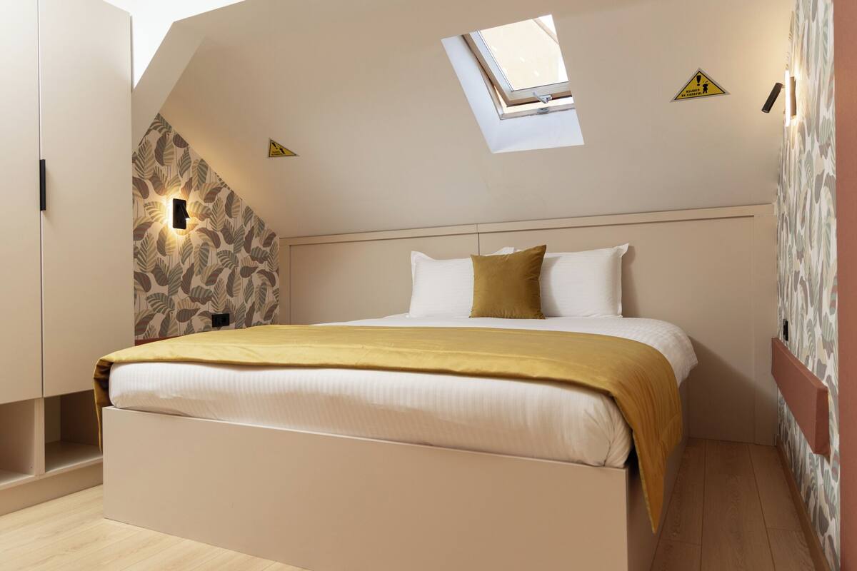 A bedroom with a sloped ceiling, a skylight, a bed with a white and gold bedspread, and a wall with a leaf-patterned wallpaper.