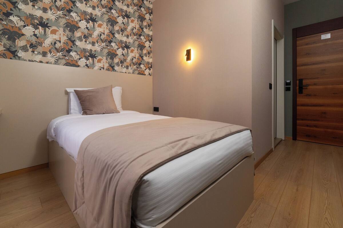 A neatly made bed with a beige blanket and pillows, a wall with a floral pattern, a wall-mounted light, and a wooden door.