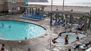 Et poolområde med liggestole og jacuzzi, udsigt til en strand og en bygning med en stenfacade.
