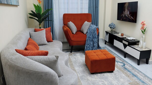 Una sala de estar con un sofá gris, un sillón naranja y un otoman a juego. Hay un aparador blanco y negro con adornos.