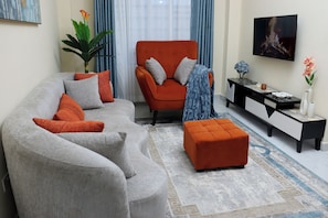 Un salon avec un canapé gris, un fauteuil orange et un pouf assorti. On y trouve également un buffet noir et blanc agrémenté d’objets décoratifs.