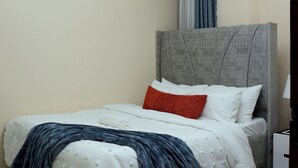 Una cama bien hecha con un cabecero gris, una alfombra gris mullida y una mesita de noche blanca con una lámpara.