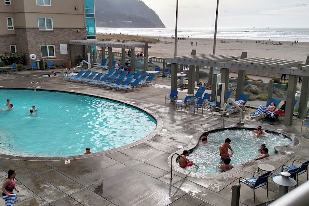 Ett poolområde med solstolar och en bubbelpool, utsikt över stranden och en byggnad med en fasad av sten.