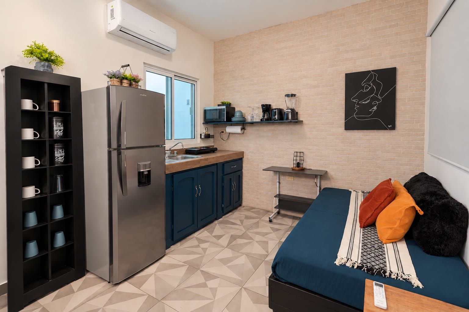 Una kitchenette compacta con refrigerador, microondas y cafetera, una cama con almohadas, y una obra de arte colgada en la pared.
