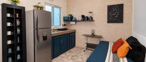 Una kitchenette compacta con refrigerador, microondas y cafetera, una cama con almohadas, y una obra de arte colgada en la pared.