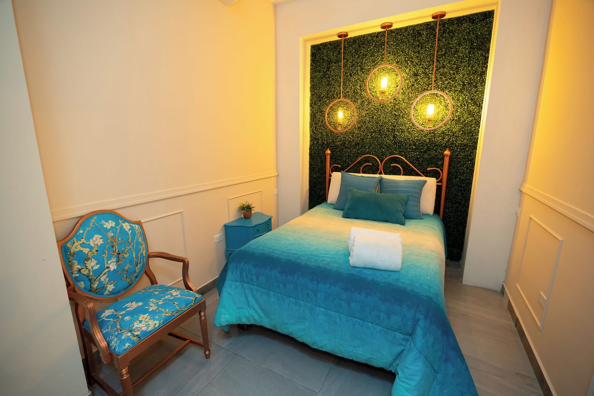 A bedroom with a bed, a chair, a nightstand, and a wall-mounted light fixture.