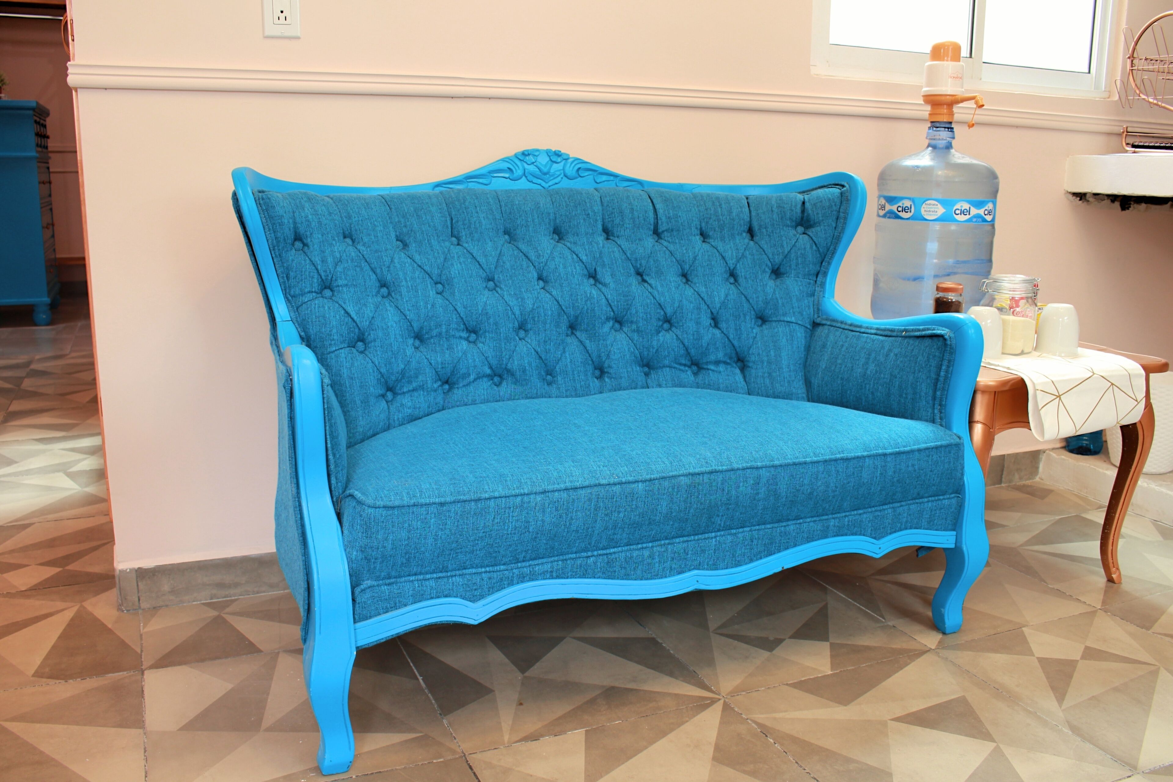 Un canapé bleu recouvert de tissu, doté d’un dossier capitonné, se trouve dans une pièce dont le sol est recouvert de carreaux décoratifs et où une fenêtre surplombe un évier.