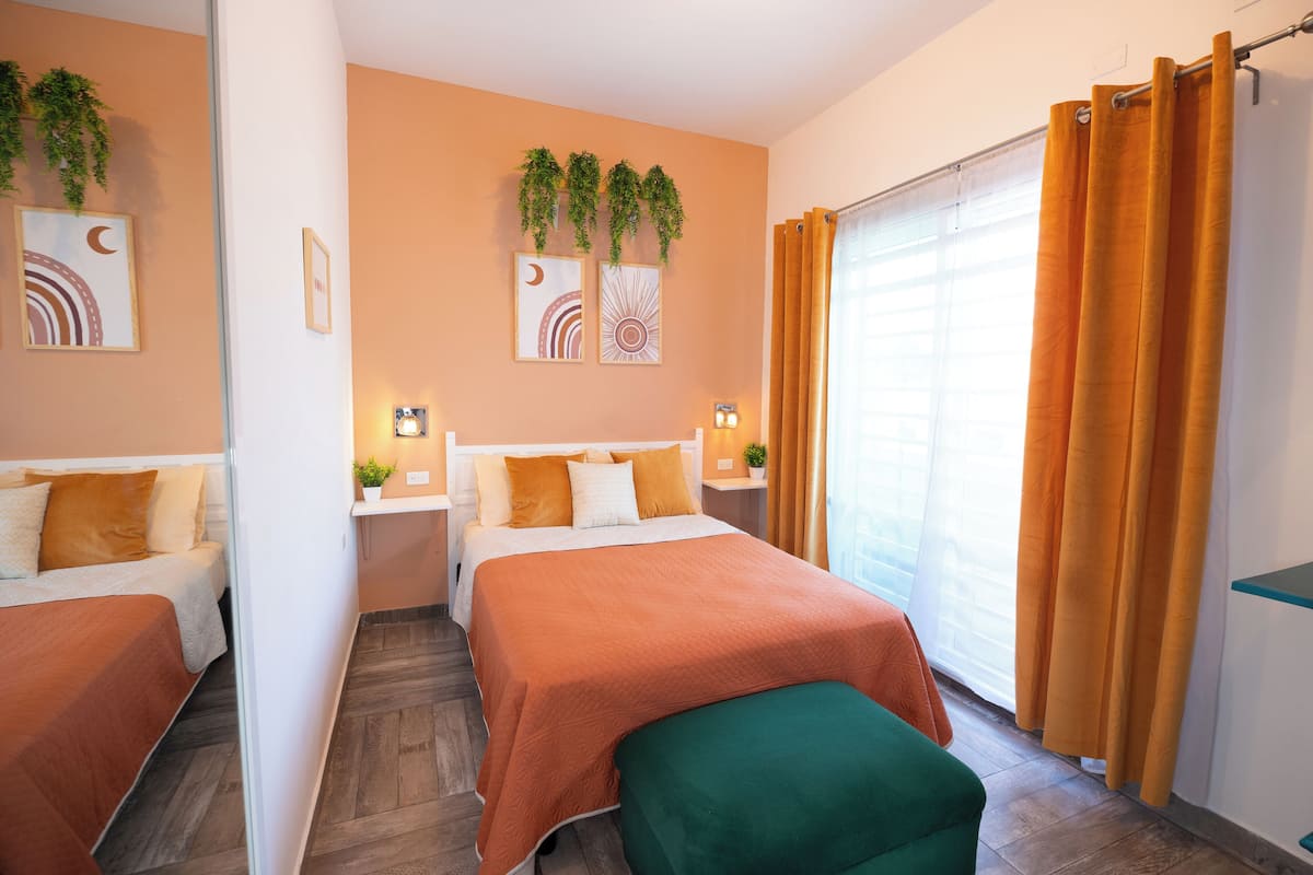 A hotel room with two beds, a green bench, a window with curtains, and wall art.