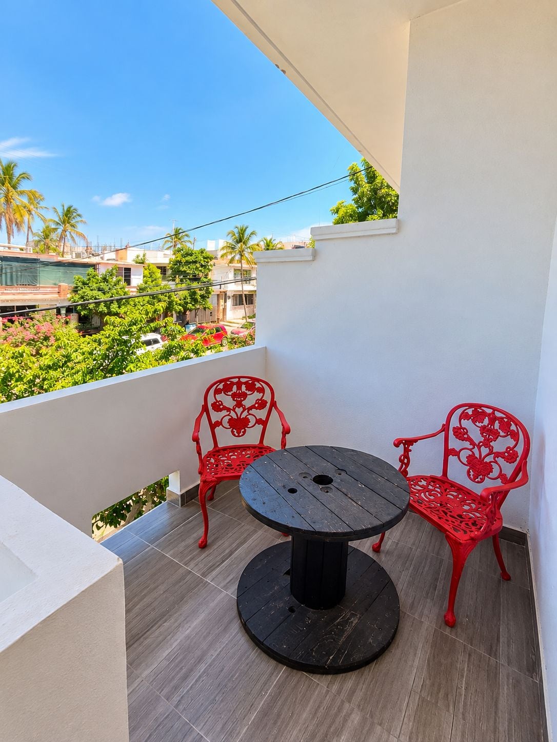 Un balcón con dos sillas rojas y una mesa redonda, con vistas a una zona residencial con palmeras y casas.