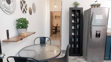 Une cuisine moderne avec une table ronde en verre, des chaises noires, un meuble intégré noir avec des étagères, un réfrigérateur en acier inoxydable et des œuvres d’art décoratives accrochées au mur.