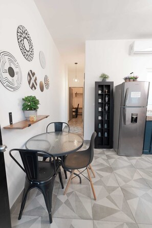 A modern kitchen with a round glass table, black chairs, a built-in black cabinet with shelves, a stainless steel refrigerator, and decorative wall art.