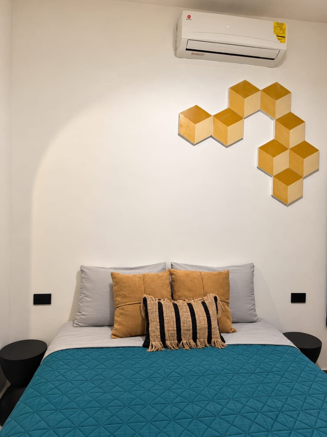 Une chambre à coucher avec un lit, des oreillers, une couvre-lit matelassée bleue, un climatiseur fixé au mur et une décoration murale aux motifs géométriques.