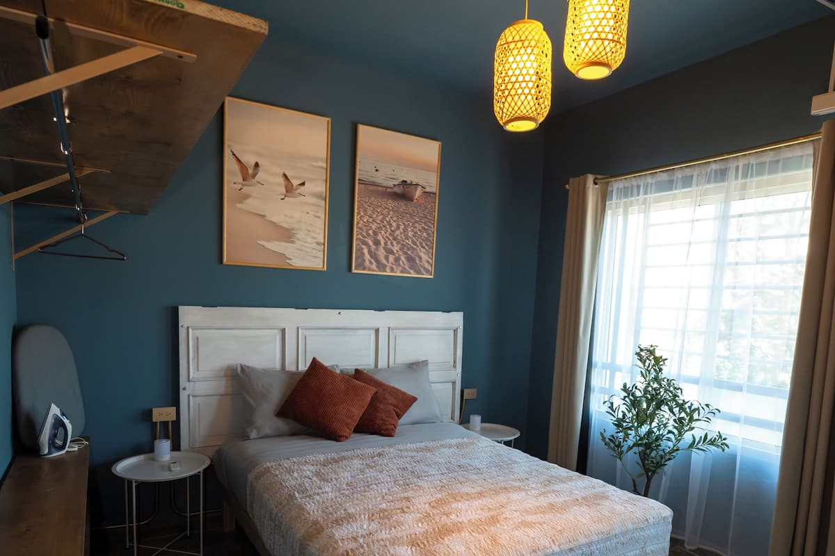 A bedroom with a large bed, a small bedside table, a hanging light fixture, and a window with curtains.