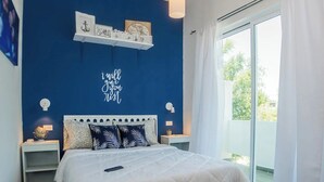 Un dormitorio con una pared azul de acento, cama con ropa blanca, dos almohadas decorativas, una mesita de noche con lámpara y una estantería con marcos.