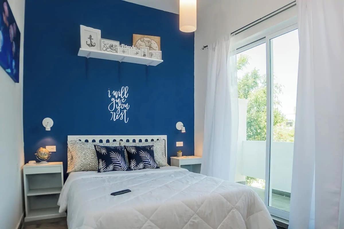 A bedroom with a blue accent wall, a bed with white bedding, two decorative pillows, a bedside table with a lamp, and a shelf with framed pictures.
