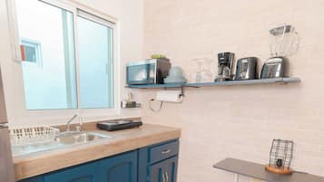 Une cuisine avec une fenêtre, un évier, une cuisinière, un meuble bleu, une étagère avec des appareils de cuisine et une petite table avec une lampe.