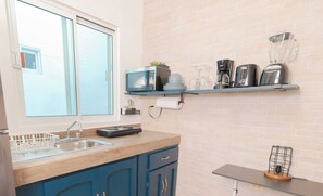 A kitchen with a window, a sink, a stove, a blue cabinet, a shelf with kitchen appliances, and a small table with a lamp. - Casa Navarro (La Paz)