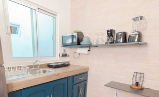 A kitchen with a window, a sink, a stove, a blue cabinet, a shelf with kitchen appliances, and a small table with a lamp.