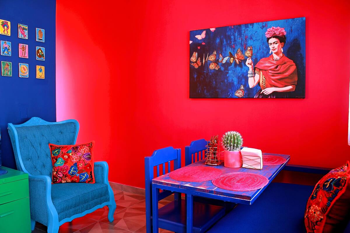 A room with a red wall, a blue sofa with a colorful cushion, a dining table with a cactus and a pink box, and a painting of a woman with butterflies.