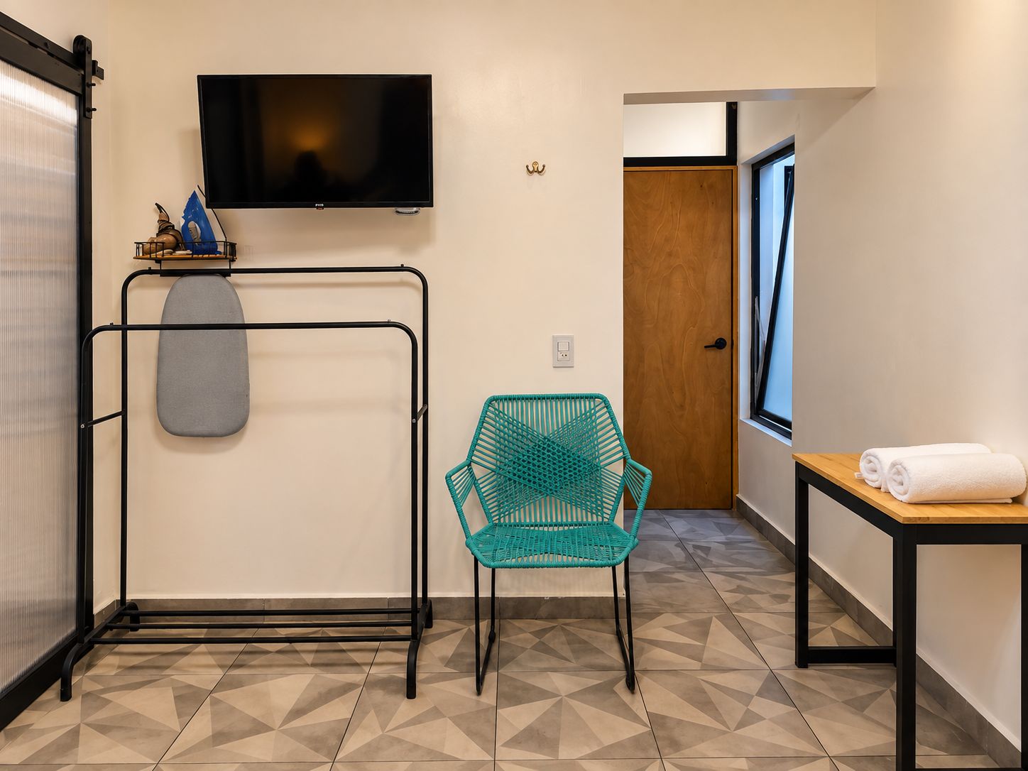 Habitación de hotel con televisor, una percha con tabla de planchar, una silla verde azulada, una mesa de madera con toallas y un suelo estampado.