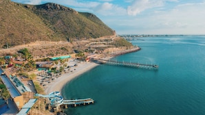 Un complejo turístico costero con playa, un muelle y montañas al fondo.