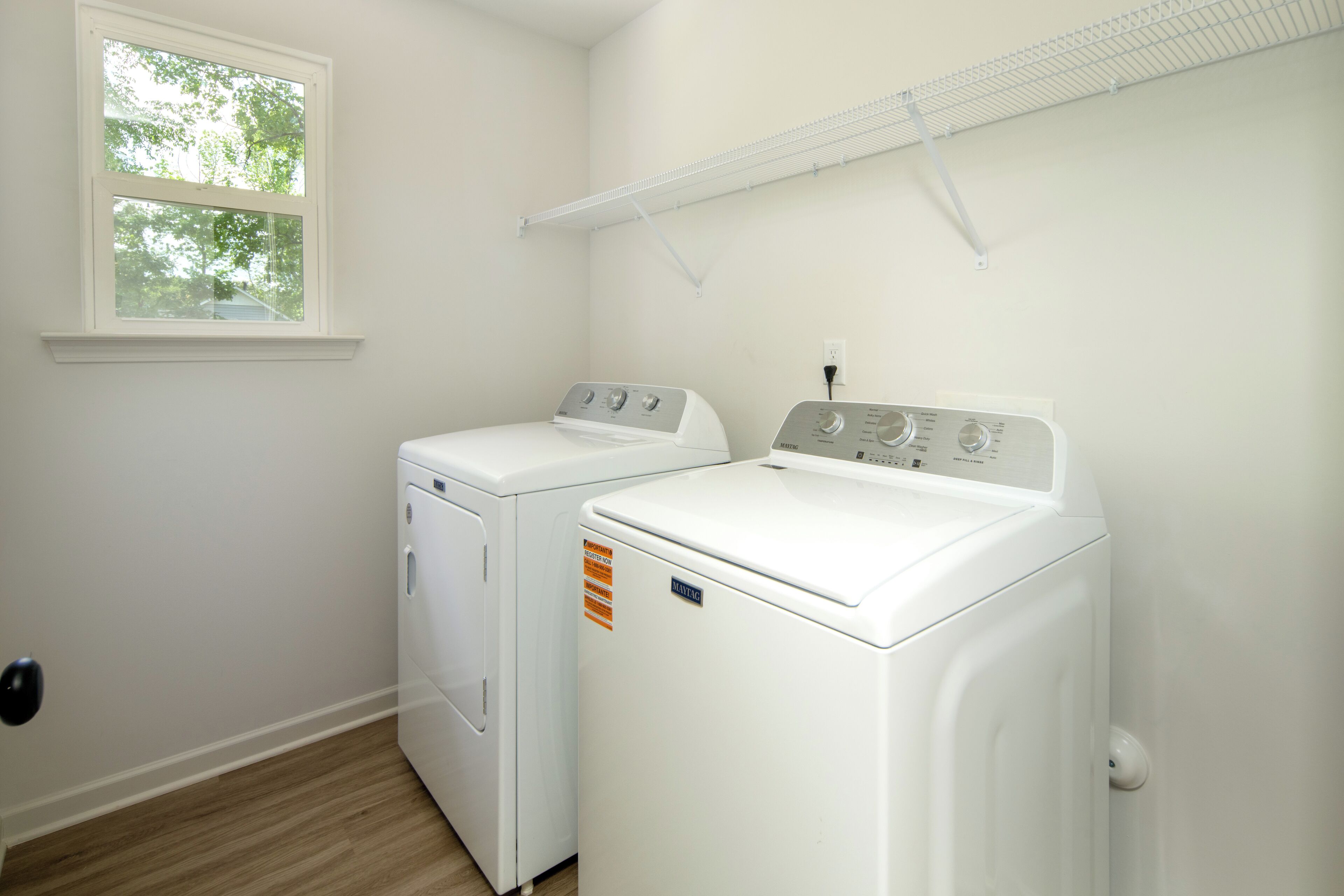 A laundry room with two white front-loading washers and dryers, a window, and a metal shelf above the dryers.