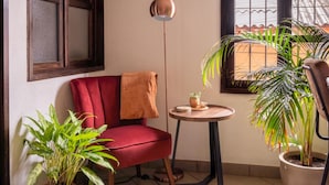 Una habitación de esquina con una silla roja, una pequeña mesa redonda con una planta, una lámpara de pie y una ventana con vistas a un edificio.