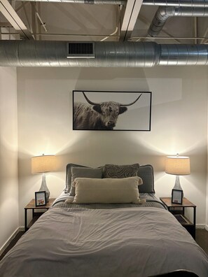 A bedroom with a bed, two bedside lamps, a framed picture on the wall, and a vent above the bed.