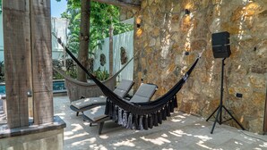Un fauteuil suspendu, des chaises longues et un haut-parleur dans un espace extérieur dont les murs sont en pierre.