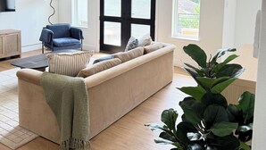 Un salon moderne avec un canapé beige, un fauteuil bleu, un ventilateur de plafond noir et une grande fenêtre donnant sur un paysage verdoyant.