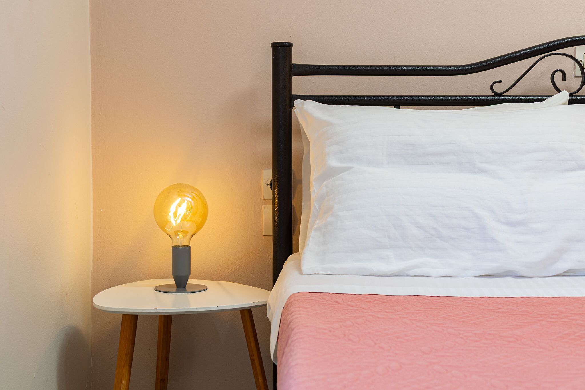 A bed with white pillows and a pink blanket, a round white nightstand with a light bulb, and a black metal headboard.