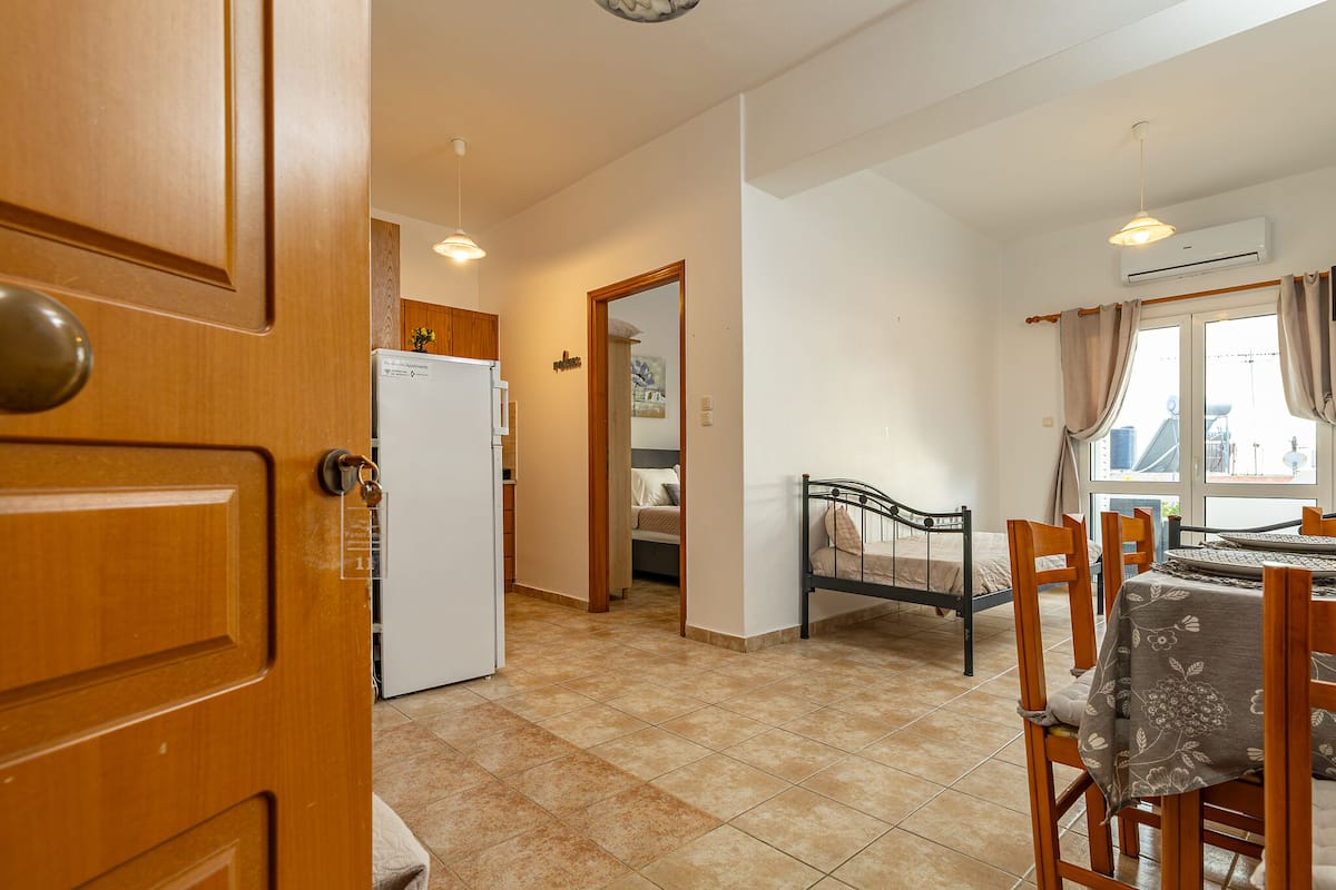 Ein Zimmer mit einem Bett, einem Esstisch mit Stühlen, einem Kühlschrank und einem Fenster mit Vorhängen.