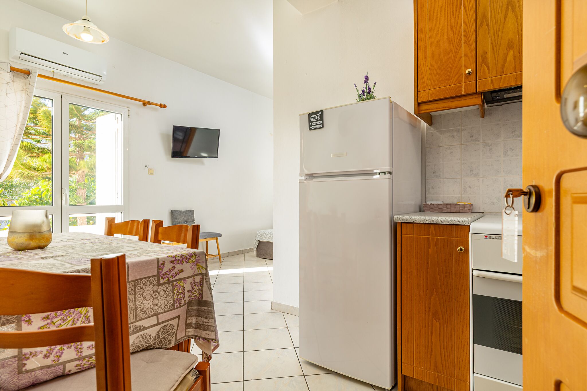 A compact kitchen with a refrigerator, oven, and wooden cabinets. A dining table with chairs is positioned near a window with a view of greenery.