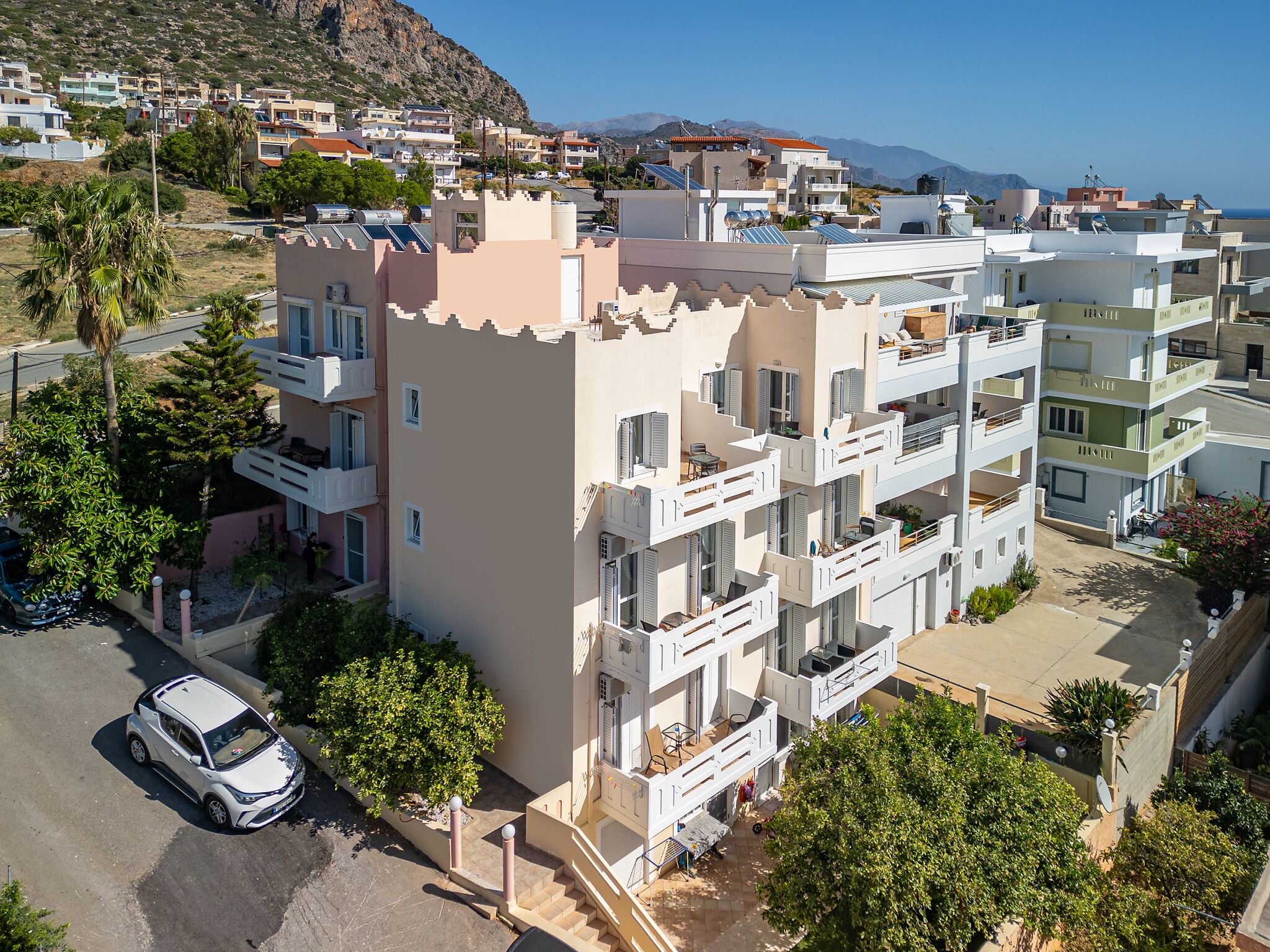 A multi-story residential building with balconies, a parking area, and a view of the sea and mountains in the background.