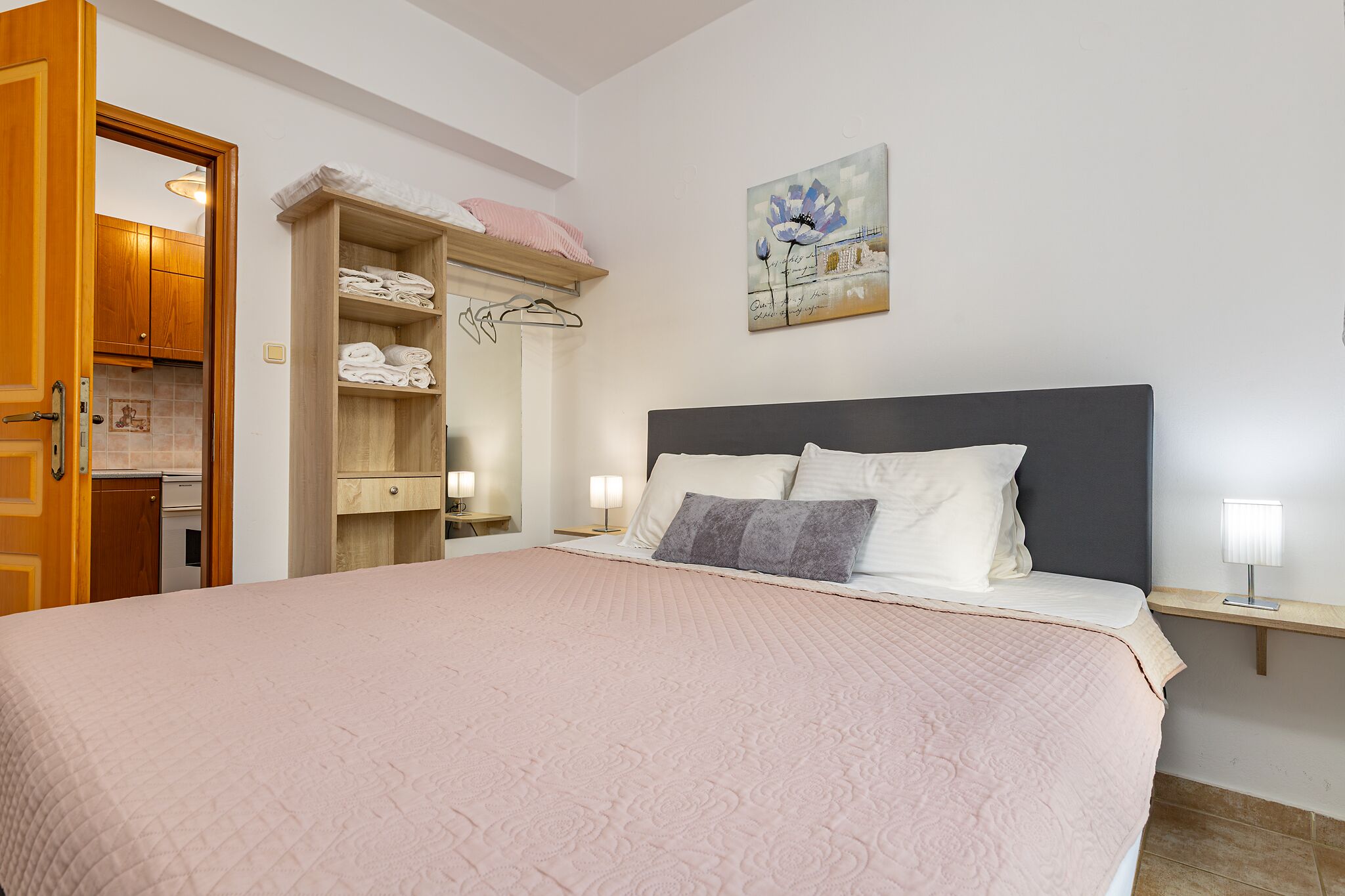 A bedroom with a large bed, a wooden wardrobe, a painting on the wall, and a bedside table with a lamp.