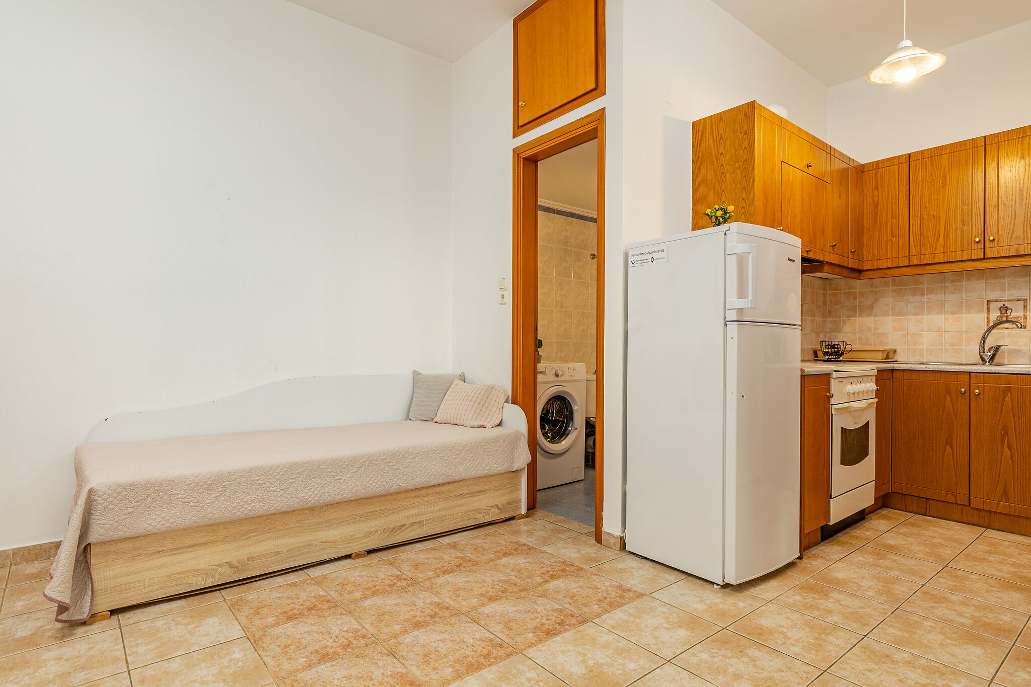 A compact room with a bed, a washing machine, a refrigerator, and wooden cabinets.