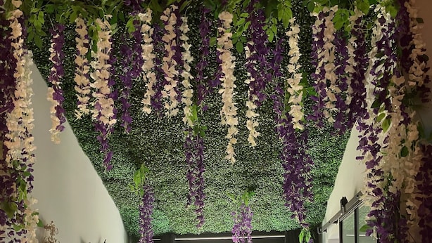 A room with a window, a table, chairs, and hanging purple and white flowers.