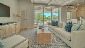 A spacious living room with a large sofa, a coffee table, and a television. There is a sliding glass door leading to a balcony with outdoor furniture. - Elegant 3-Bed, 3-Bath Penthouse with Panoramic Ocean Views at Watercolors (Roatán)