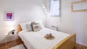 Ein Schlafzimmer mit Bett, Nachttisch, Lampe, gerahmtem Bild, Fenster und Spiegel an der Wand.