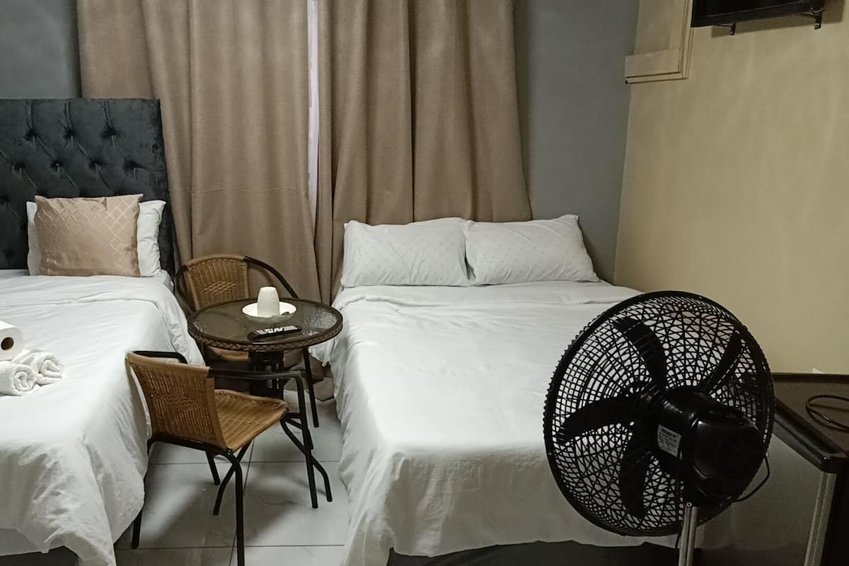 A hotel room with two beds, a small table with a chair, a fan, and a towel on the bed.