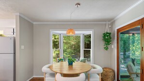 A dining area with a round wooden table, white furry chairs, a hanging orange pendant light, and a view of greenery through large windows.