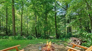 A forested area with a fire pit and two wooden chairs.