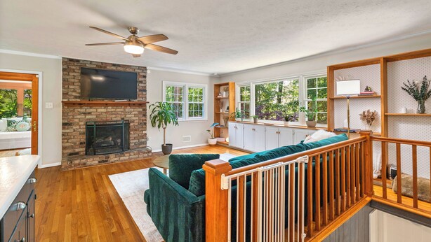 A spacious living room with a fireplace, a ceiling fan, and a large window overlooking greenery.